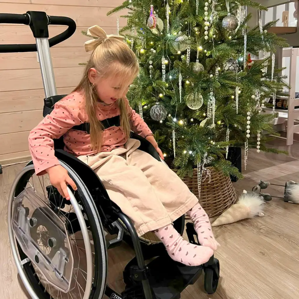 Mergaitė vežimėlyje prie papuoštos Kalėdų eglutės